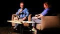 A white man sits in a chair holding a microphone opposite a white woman