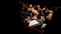 An audience from the back in red velvet chairs.