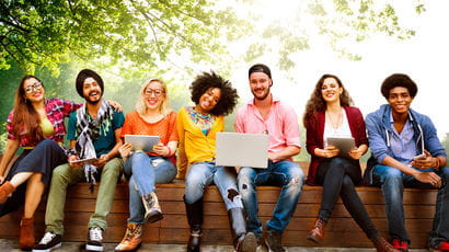 A group of students sitting outside and smiling at the camera, whilst holding different types of mobile devices (laptops, tablets, phones).