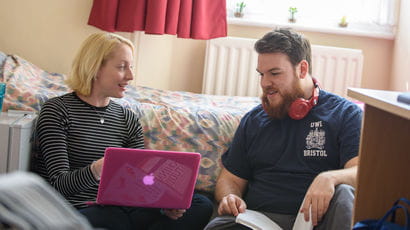Two students are sitting in student accommodation, using a laptop and a textbook.